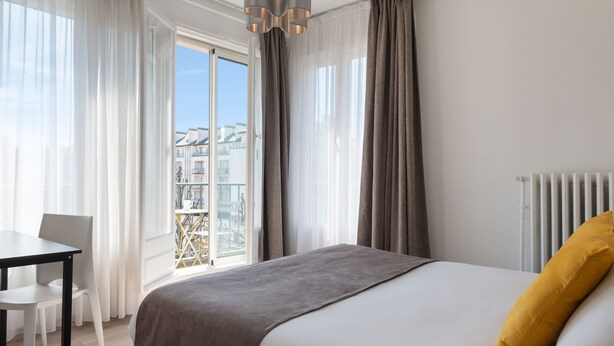Een moderne hotelkamer met een groot bed, uitzicht op de stad vanaf een balkon, een eettafel en een radiator.