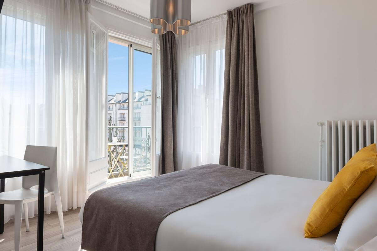 Une chambre d’hôtel moderne avec un grand lit, une vue sur la ville depuis un balcon, une table à manger et un radiateur.