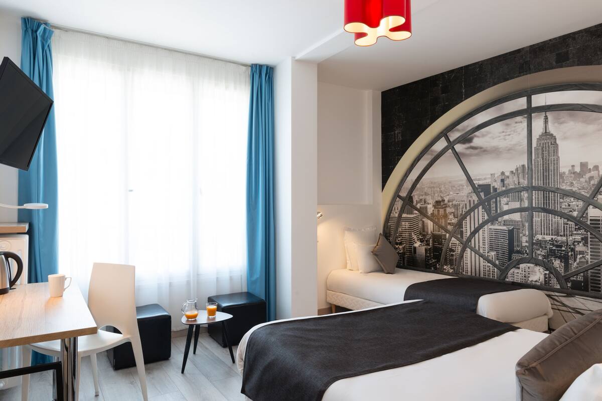 Une chambre d’hôtel avec une fresque représentant un paysage urbain, un lit, un bureau avec une chaise, une table basse et un téléviseur fixé au mur.