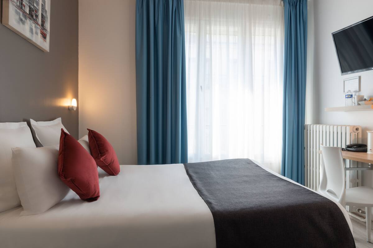 Une chambre d’hôtel avec un lit, deux oreillers rouges, un plaid noir, un bureau blanc avec un ordinateur et un téléviseur fixé au mur.