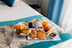 Un plateau de petit-déjeuner avec des croissants, des œufs, des crêpes, du jus de fruits et un petit bol de chocolat, le tout posé sur une table de chevet.