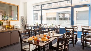 Een eetruimte met meerdere tafels en stoelen, ontbijtartikelen zoals croissants en sinaasappelsap, en uitzicht op de straat.