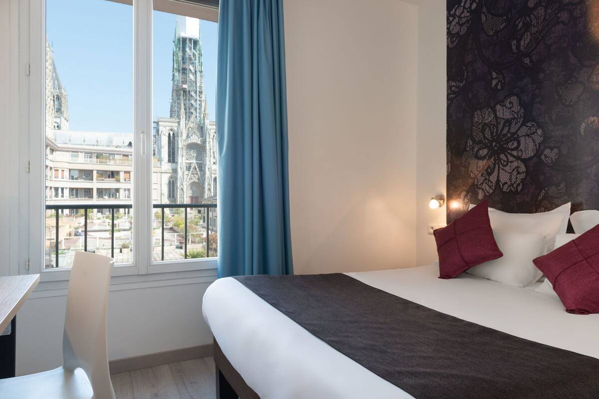 Une chambre d’hôtel avec un lit, un bureau, une chaise, une fenêtre donnant sur un immeuble et un mur décoré d’un motif.
