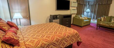 A hotel room with a large bed, a desk with a computer, a TV, and two armchairs.