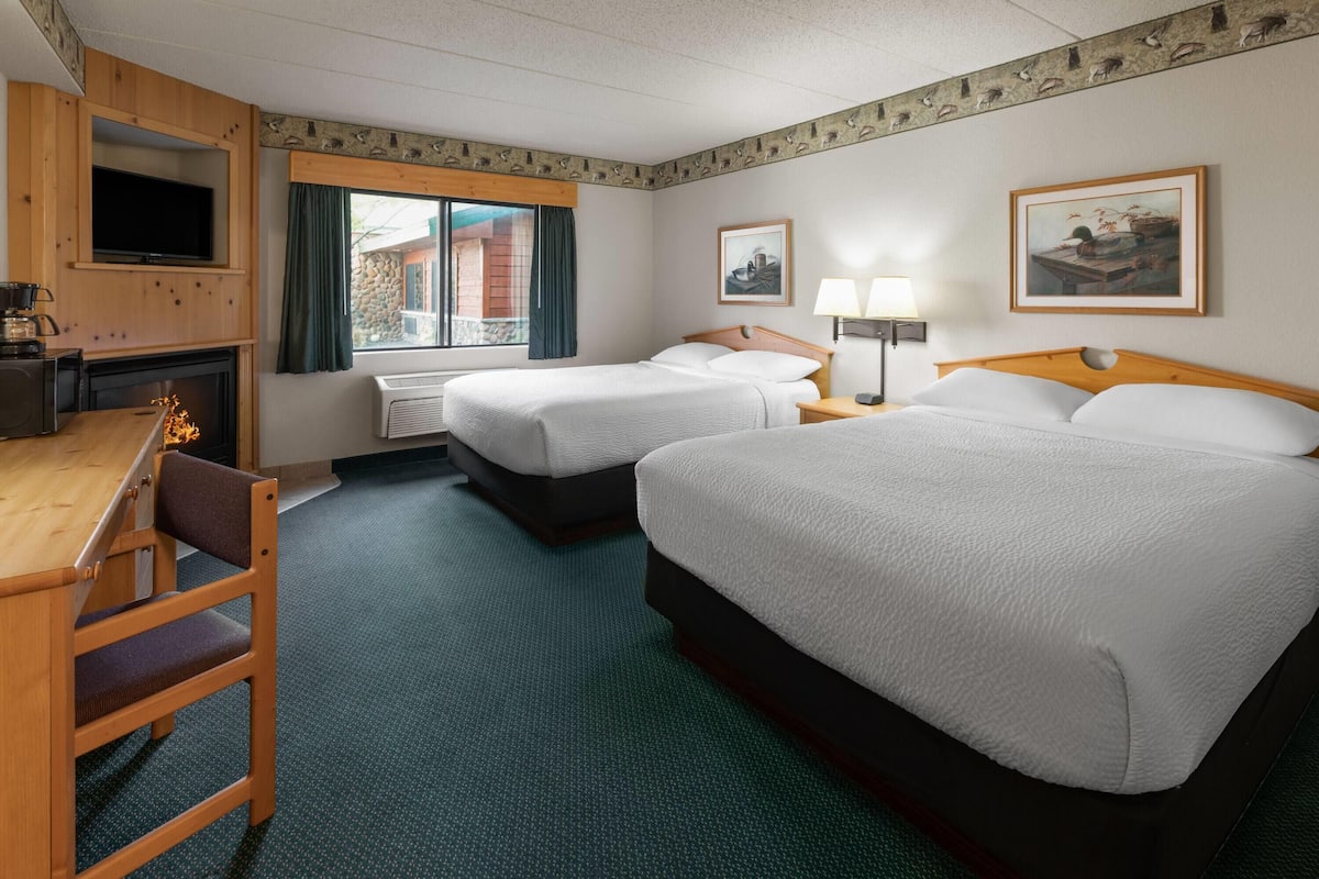 Quarto de hotel com duas camas, uma escrivaninha com cadeira, lareira, televisão e obras de arte nas paredes.