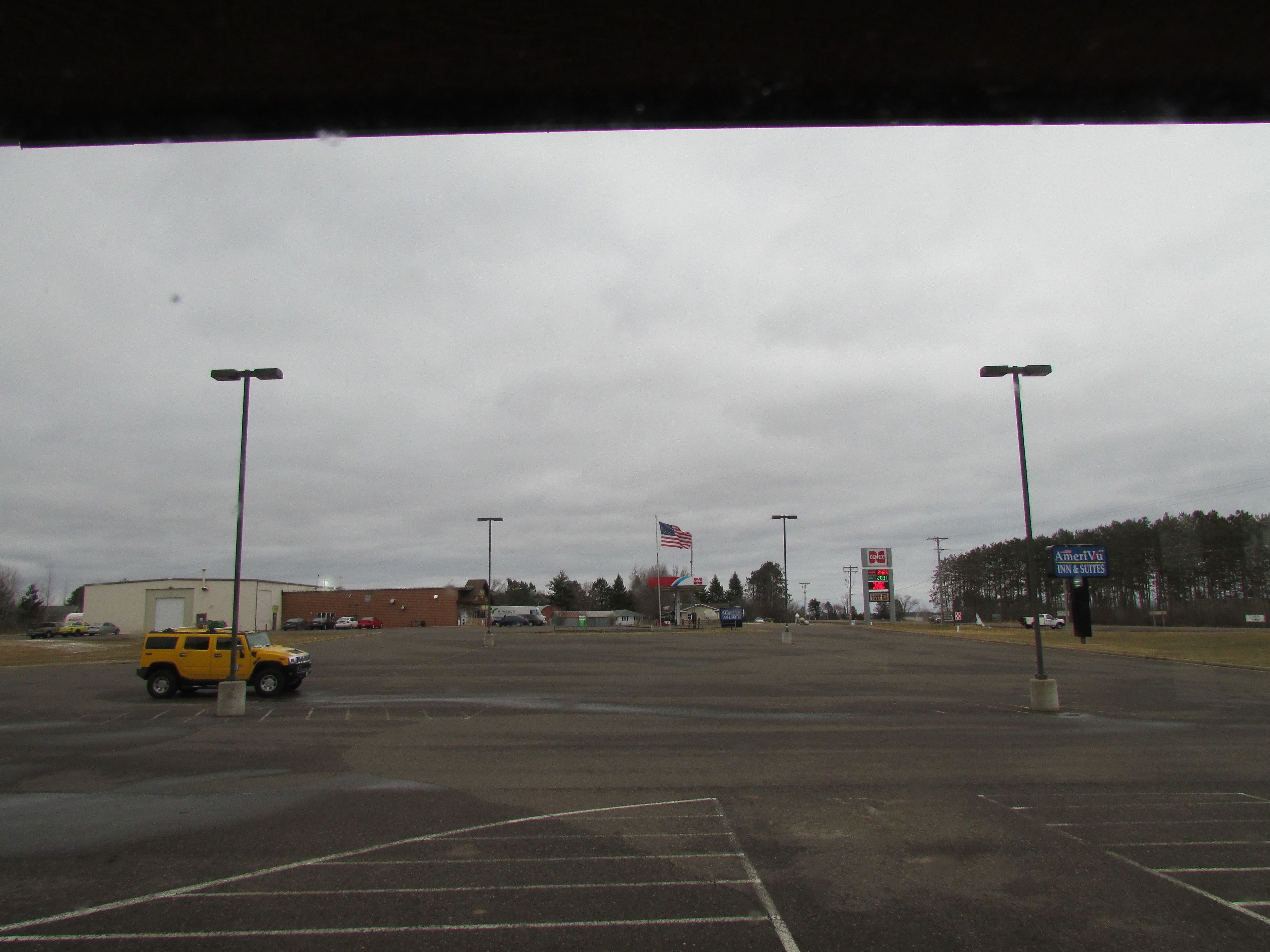 An empty parking lot with a yellow SUV, a gas station, and a flagpole with an American flag.
