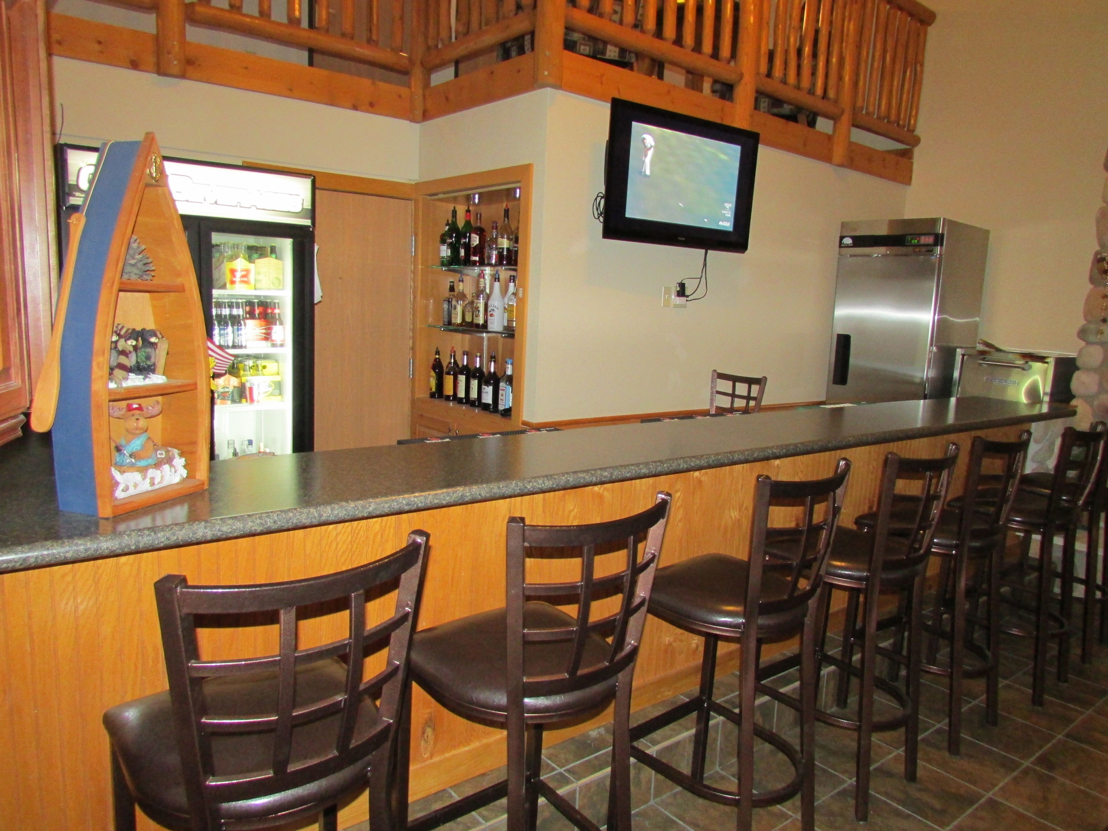 Una zona de bar con sillas de madera, un televisor montado en la pared y un refrigerador.