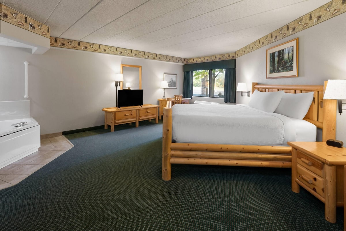 Quarto de hotel com uma cama grande, mesas de cabeceira de madeira, escrivaninha, televisão e janela com cortinas.