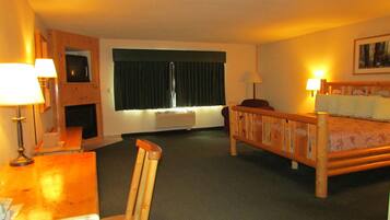 Una habitación de hotel con una cama de madera, un escritorio con teléfono, una silla, una ventana con cortinas y un calentador fijado a la pared.