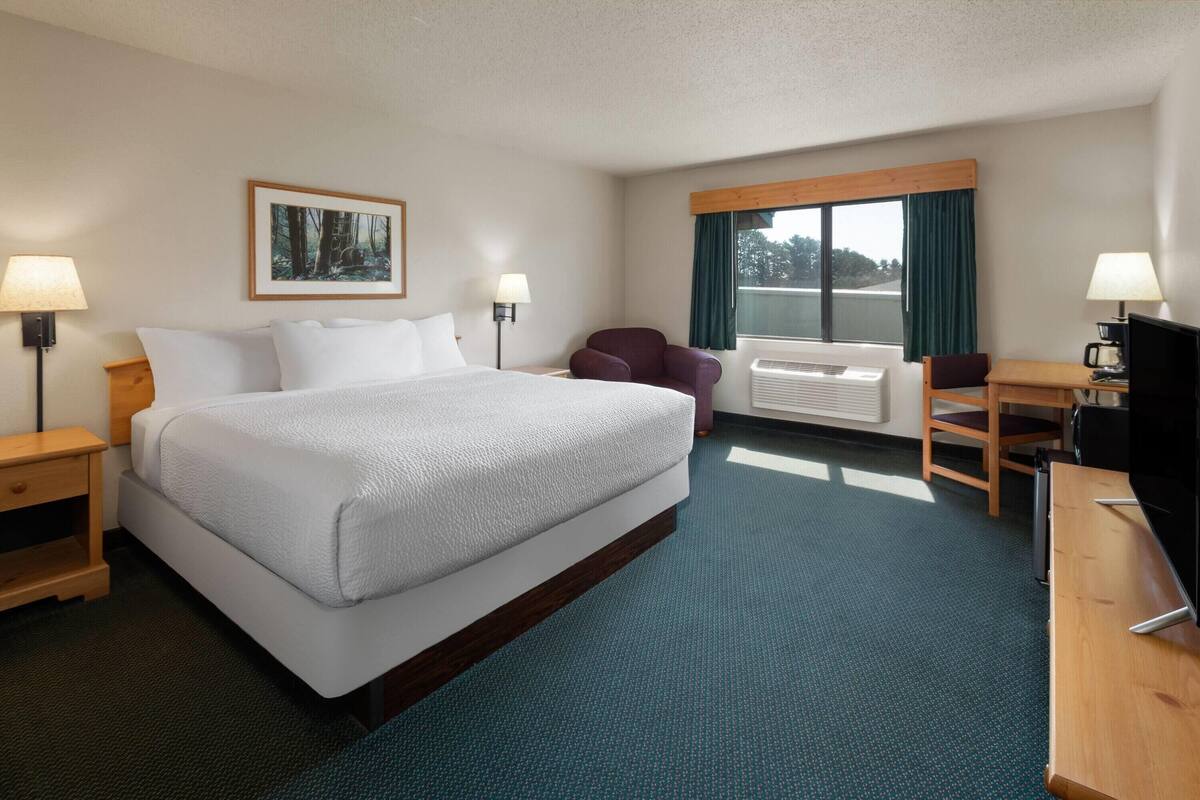 Quarto de hotel com uma cama grande, duas mesas de cabeceira com abajures, uma escrivaninha com cadeira, televisão e uma janela com cortinas.