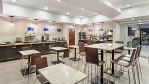 Ein moderner Hotel-Speisesaal mit mehreren Tischen und Stühlen, ein Küchenbereich mit Kaffeeautomaten und eine Theke mit einer Kaffeestation.