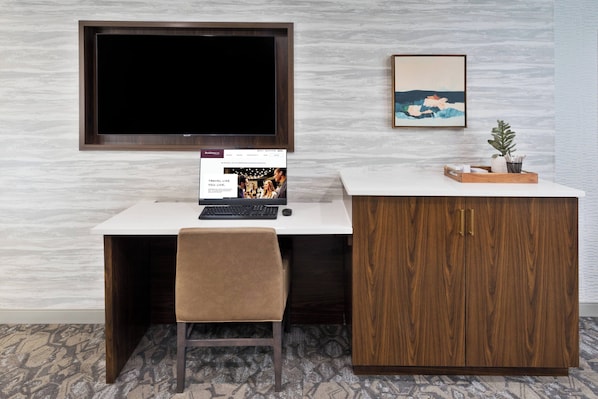 A modern hotel room with a large flat-screen TV mounted on the wall, a desk with a computer, a chair, a wooden cabinet, and a framed artwork on the wall.
