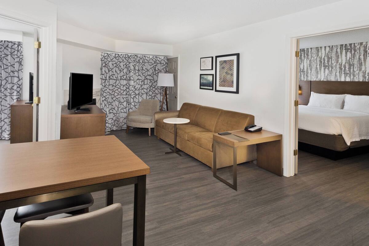 A hotel room with a wooden table, a sofa, a TV, a lamp, a bed, and framed pictures on the wall.