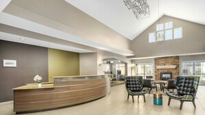 A reception area with a curved wooden counter, a small plant, and a sign that reads "MANAGEMENT BONVY". There are two black leather chairs with a plaid pattern and a round table with chairs. The room has a high ceiling with a geometric light fixture and a stone fireplace. - Residence Inn By Marriott Las Vegas Stadium Area  (Las Vegas)