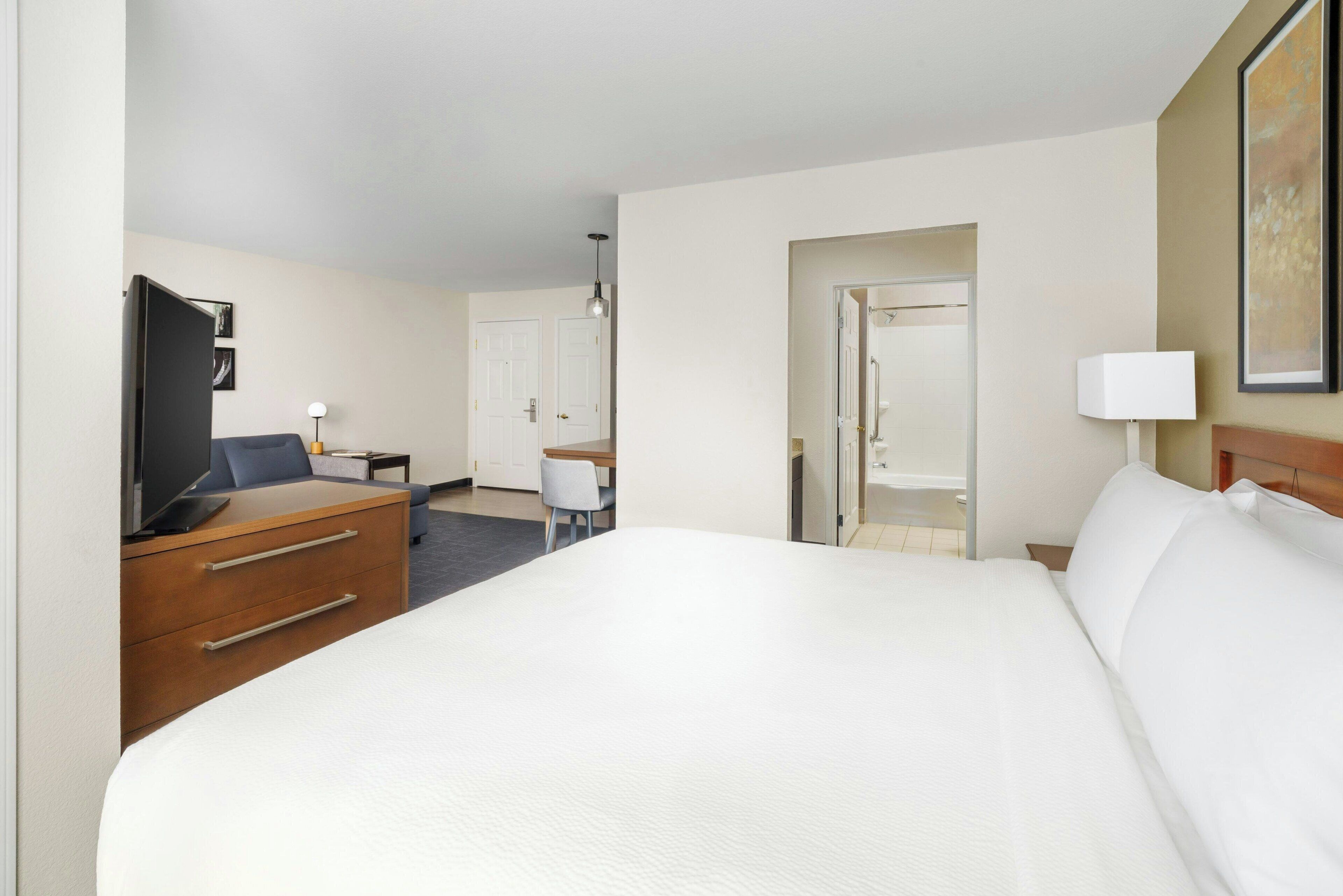 A hotel room with a large bed, a desk with a chair, a television, and a door leading to another room.