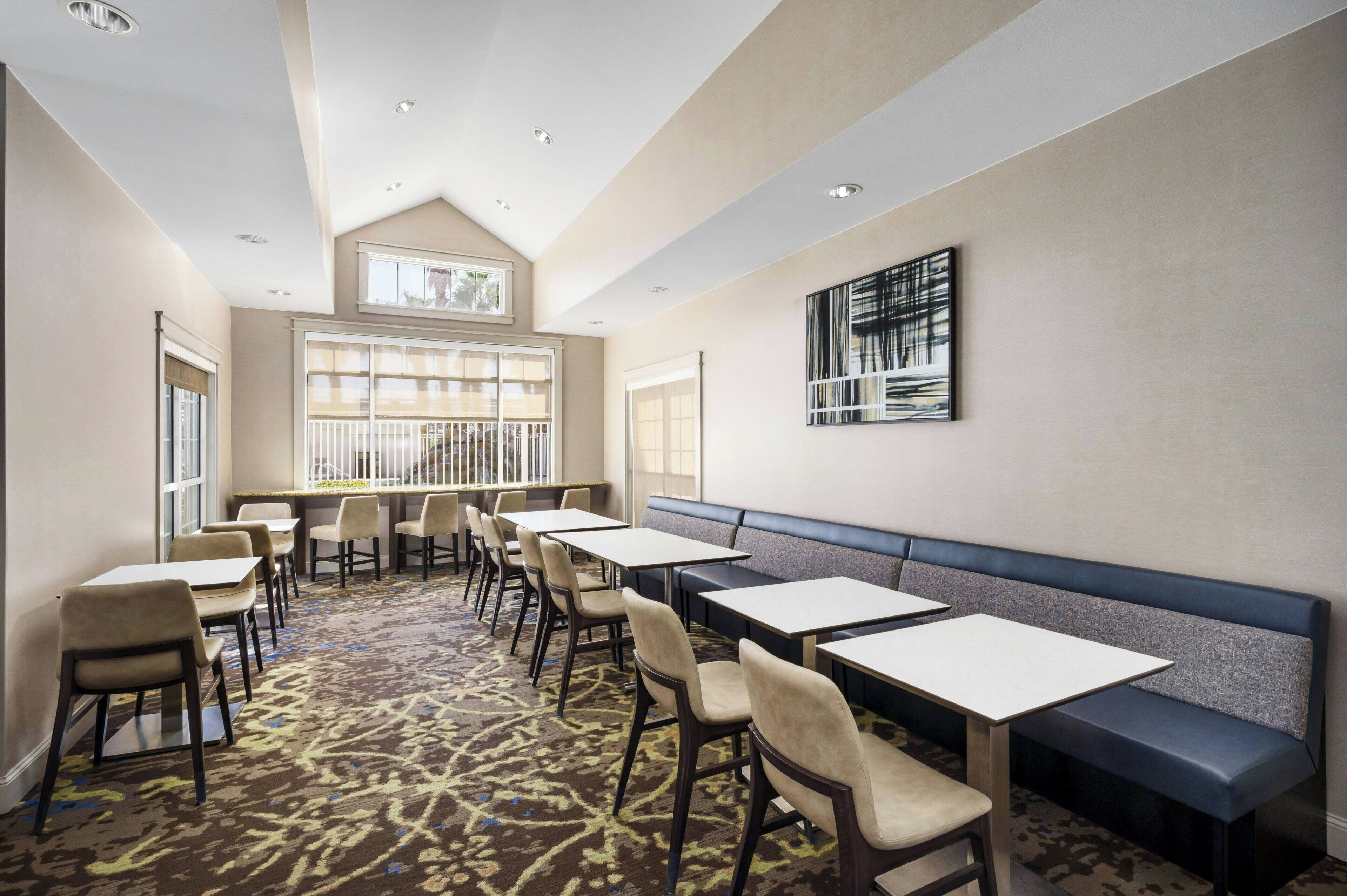 A hotel dining area with tables and chairs, a bench, and a large window with blinds.