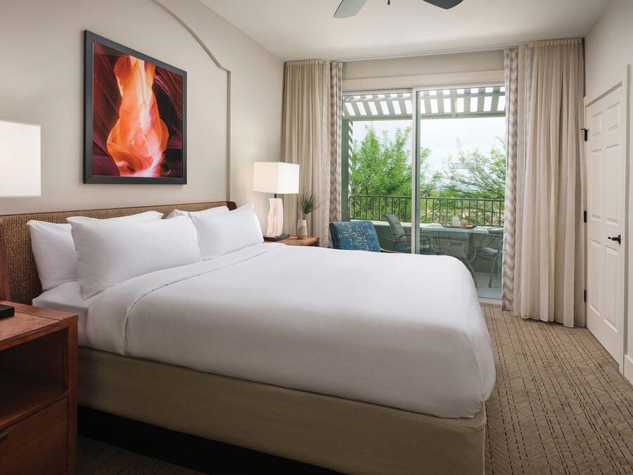 A hotel room with a large bed, a nightstand, a lamp, a window with curtains, and a view of greenery outside.