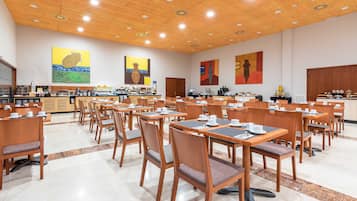 Un grand espace dédié à la salle à manger, avec des tables et des chaises en bois, une cuisine à l’arrière-plan et des œuvres d’art accrochées aux murs.
