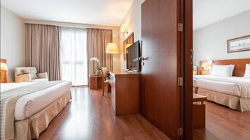 Habitación de hotel con dos camas, un escritorio, una televisión y un ventanal con cortinas.