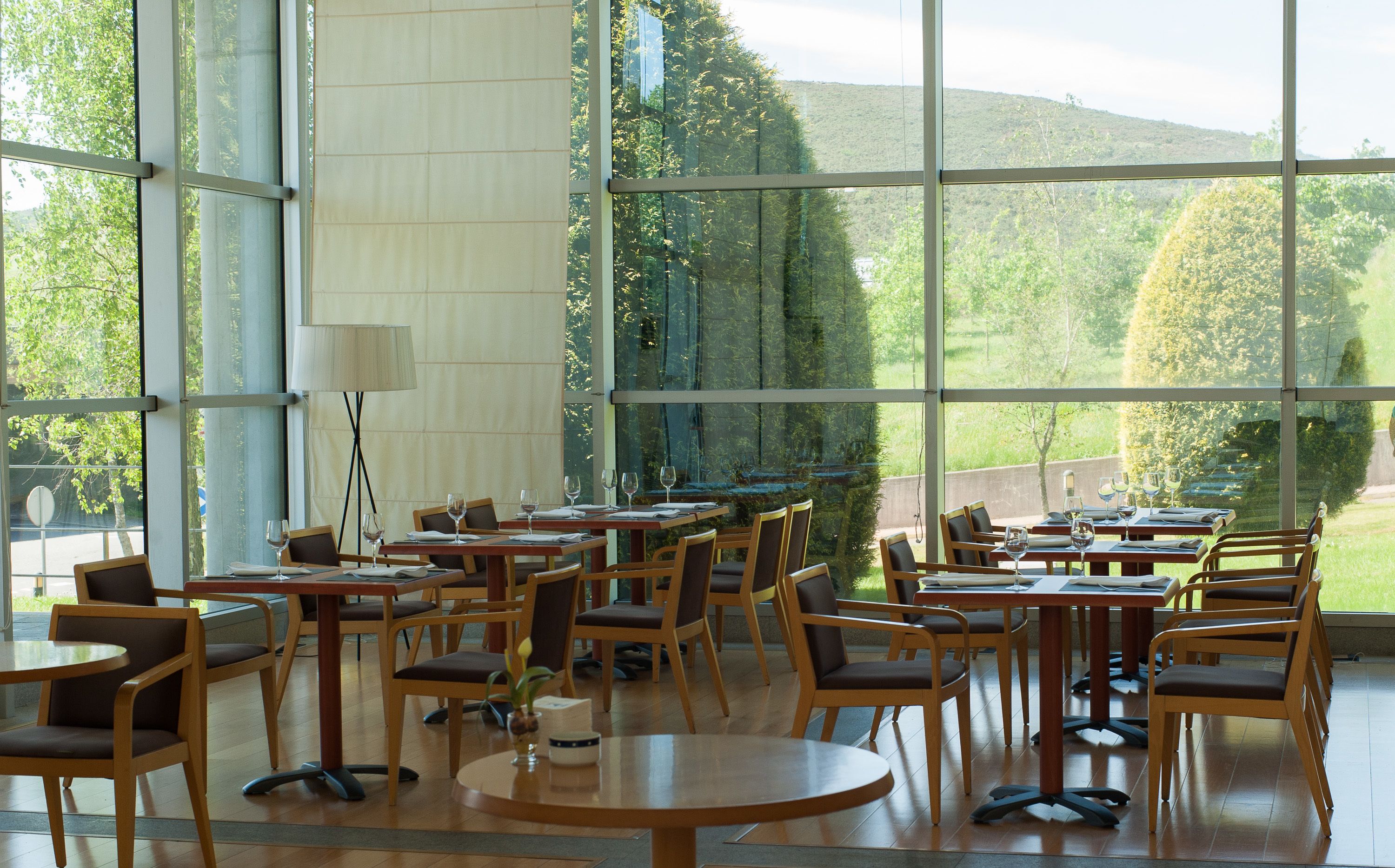 A spacious indoor dining area with large windows offering a view of a lush green landscape and hills. Tables are arranged for dining, and there are chairs with cushions.
