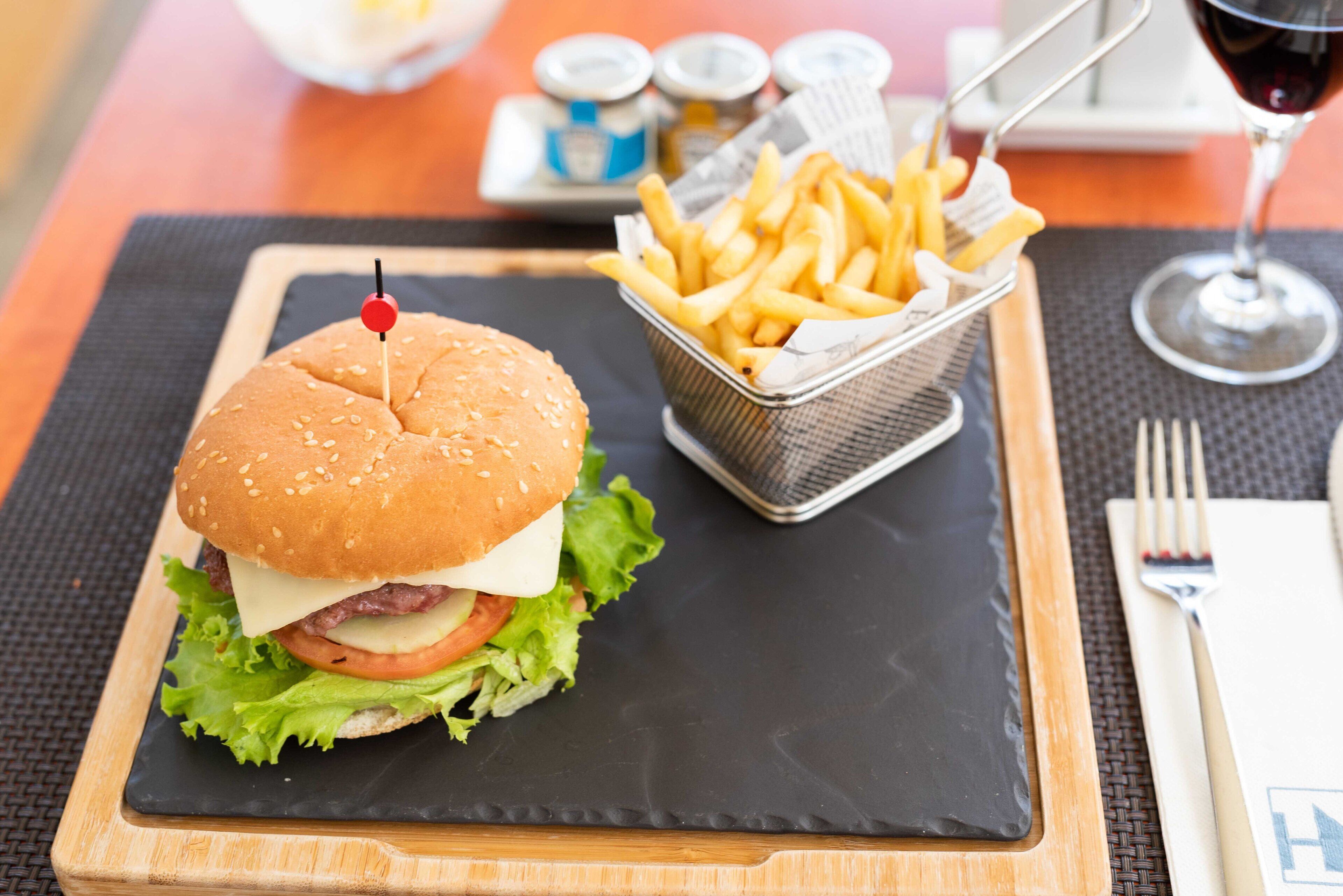 A burger with a sesame seed bun, lettuce, tomato, cheese, and a red pick on top, served on a black slate plate.