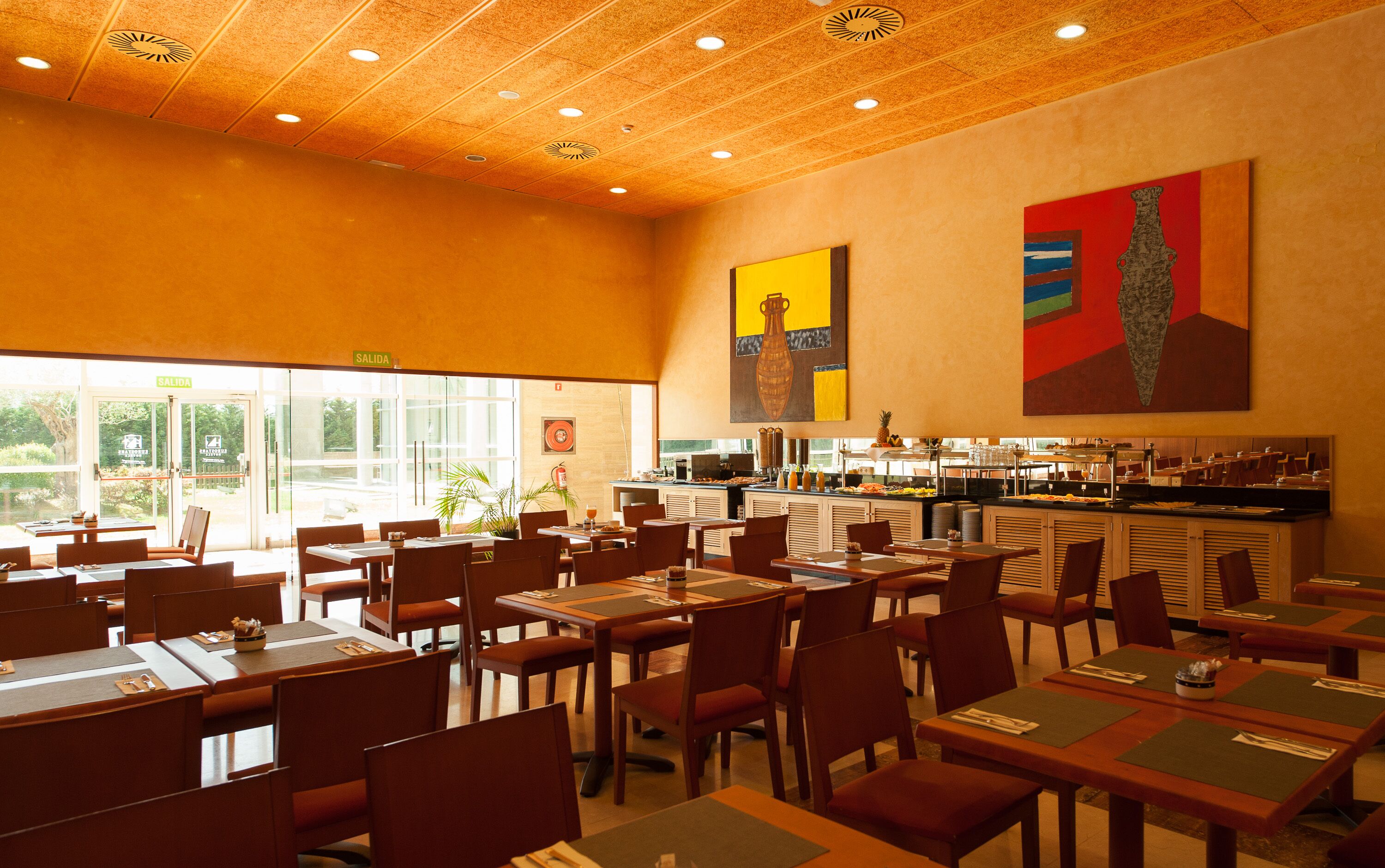 Ein Restaurant mit mehreren Tischen und Stühlen, einer Küchenzeile und großen Fenstern, die für viel Tageslicht sorgen.