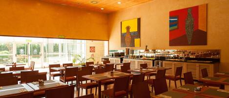 Ein Restaurant mit mehreren Tischen und Stühlen, einer Küchenzeile und großen Fenstern, die für viel Tageslicht sorgen.