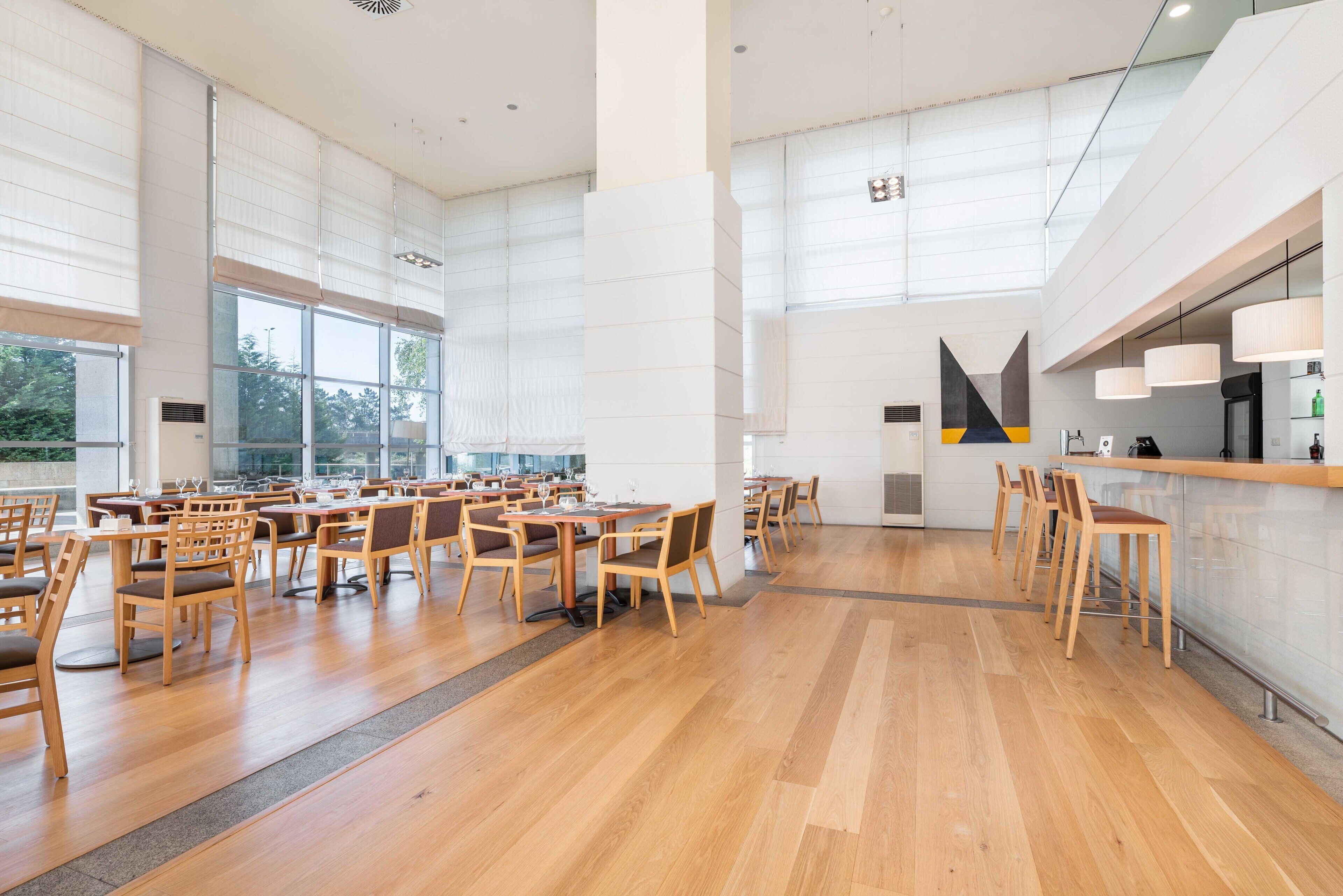 A spacious dining area with wooden tables and chairs, large windows, and a modern kitchen area.