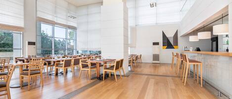 A spacious dining area with wooden tables and chairs, large windows, and a modern kitchen area.