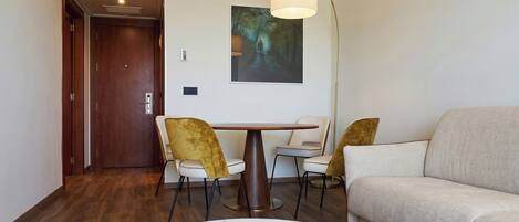 A hotel room with a wooden door, a round marble table, two chairs, a sofa, and a painting on the wall.