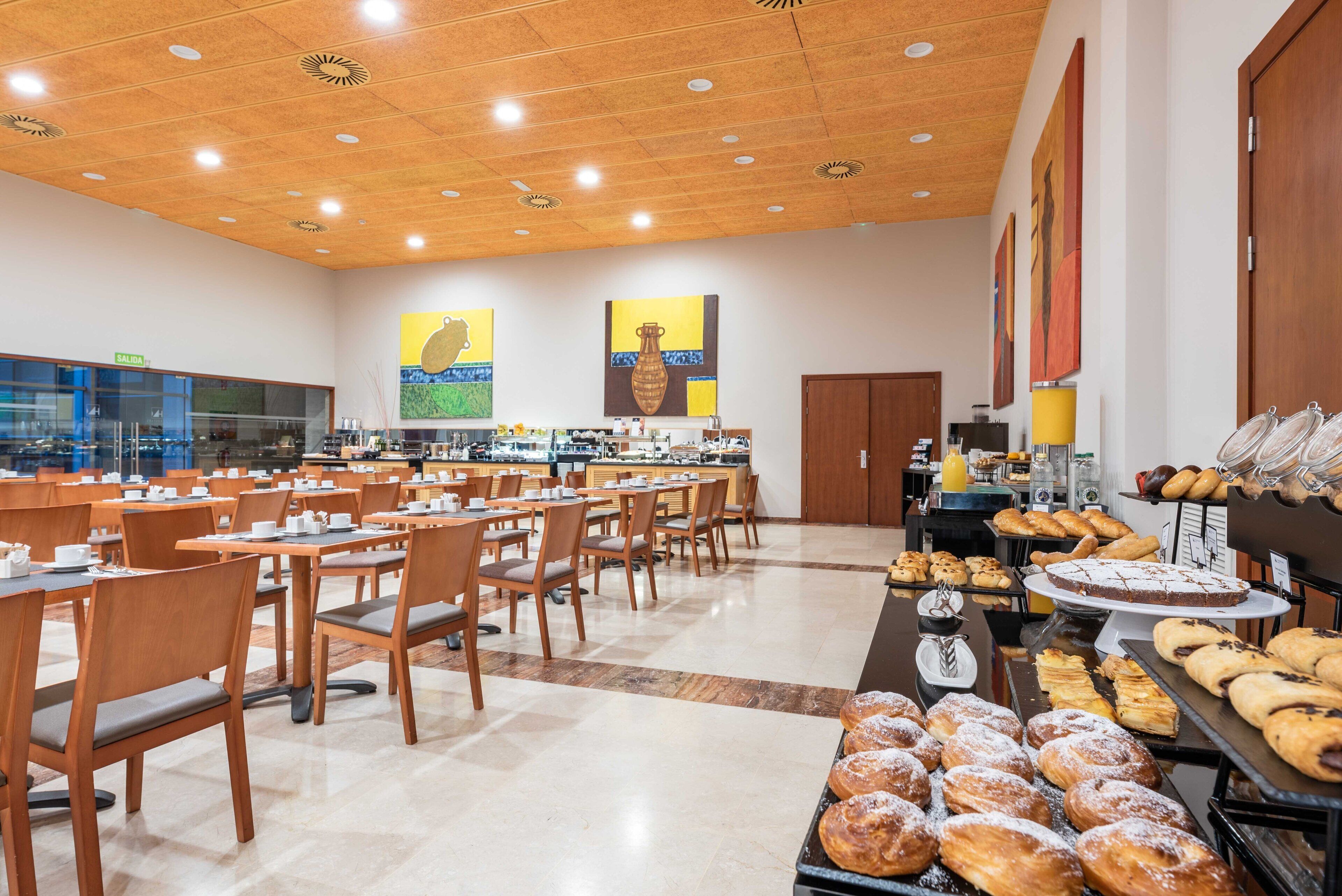 Een ruime cafetaria met meerdere tafels en stoelen, diverse gebakjes uitgestald op toonbanken, en een keuken in de achtergrond.