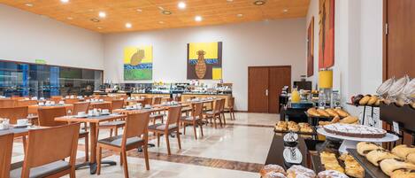 A spacious cafeteria with multiple tables and chairs, a variety of baked goods displayed on counters, and a kitchen area in the background.