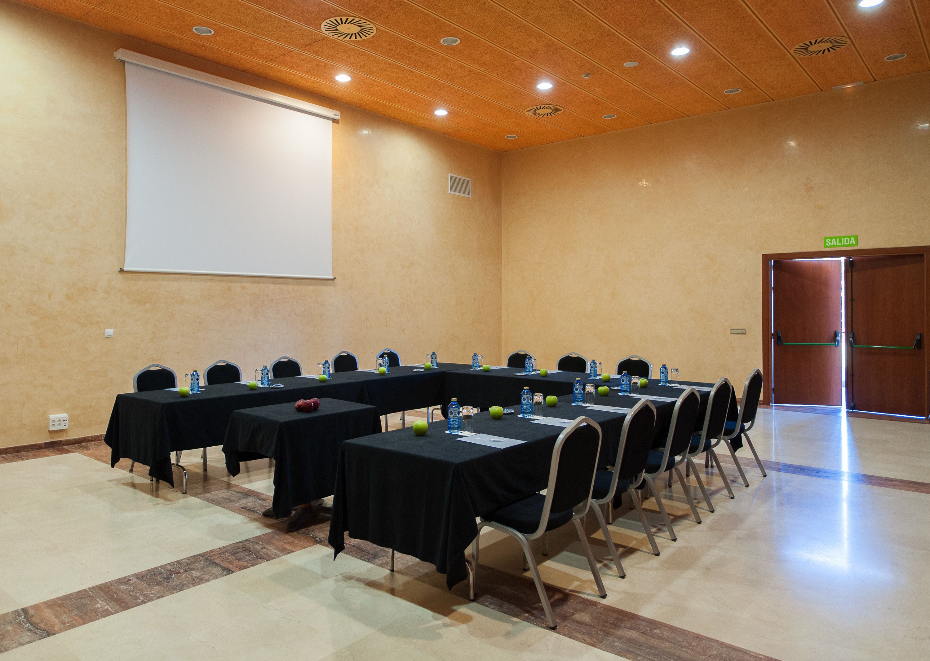 A conference room with multiple tables, chairs, a projector screen, and a door marked "SALIDA".