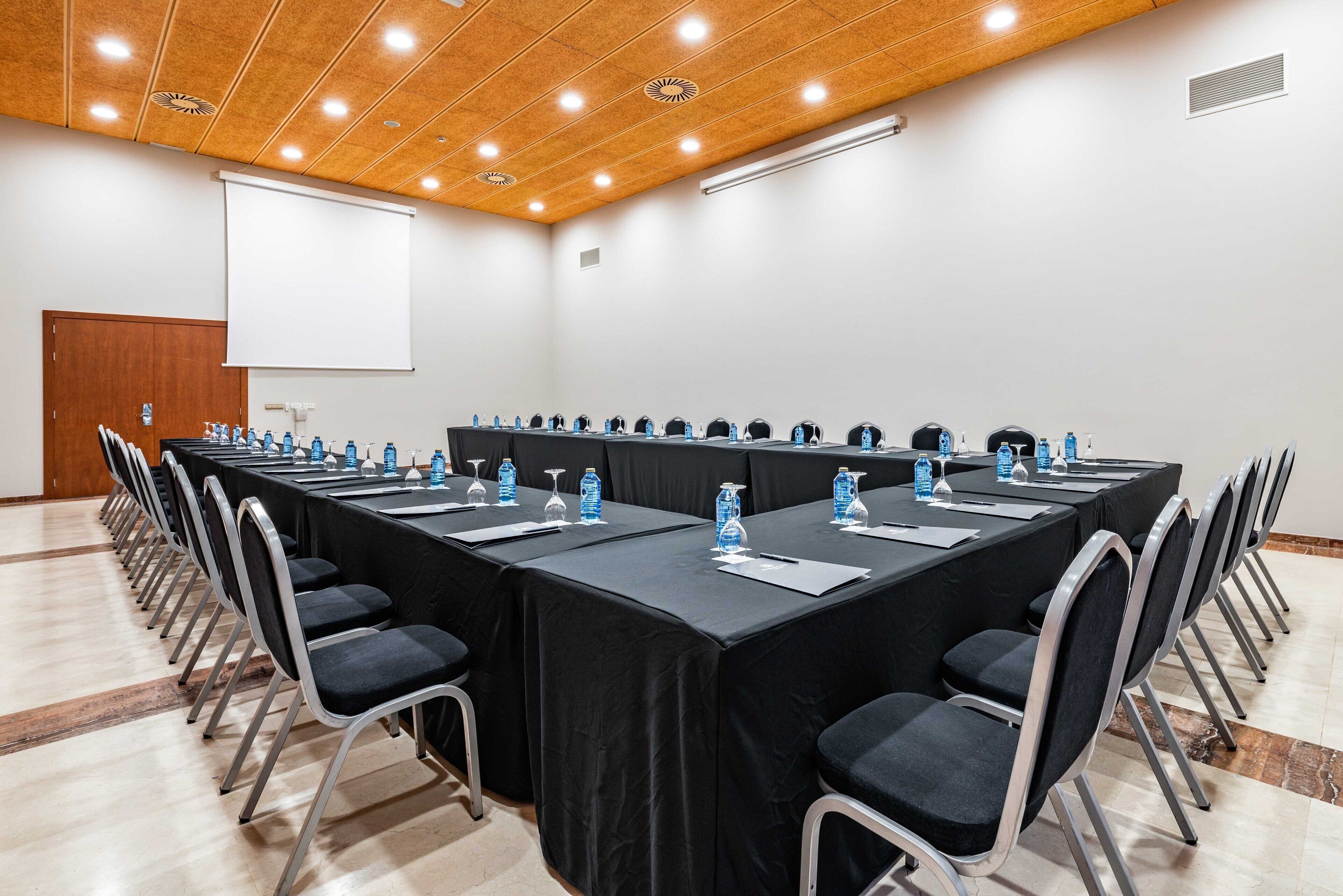 A conference room with a long table, chairs, water bottles, and a projector screen.