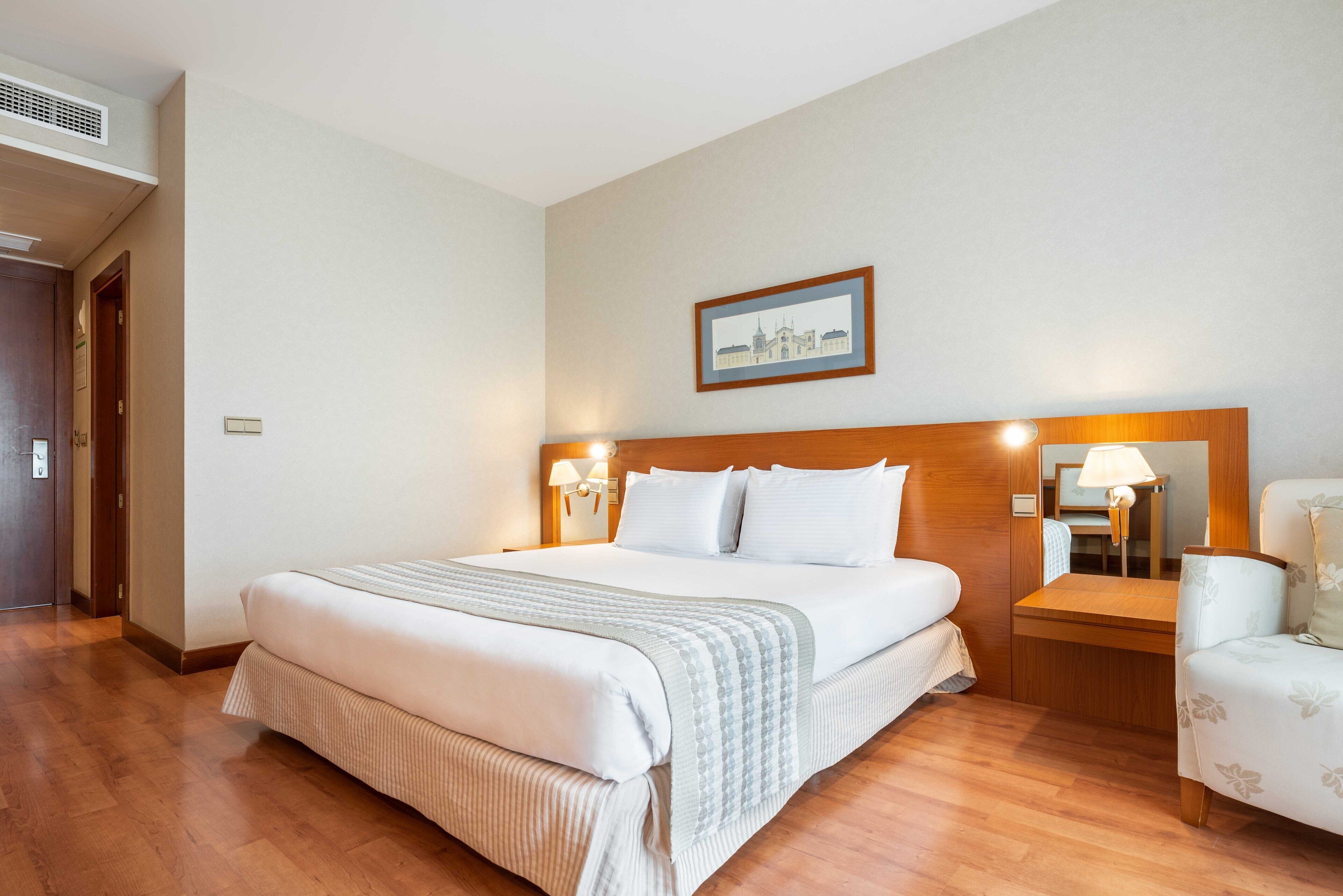 A hotel room with a large bed, wooden headboard, bedside tables with lamps, a framed picture on the wall, and a wooden floor.