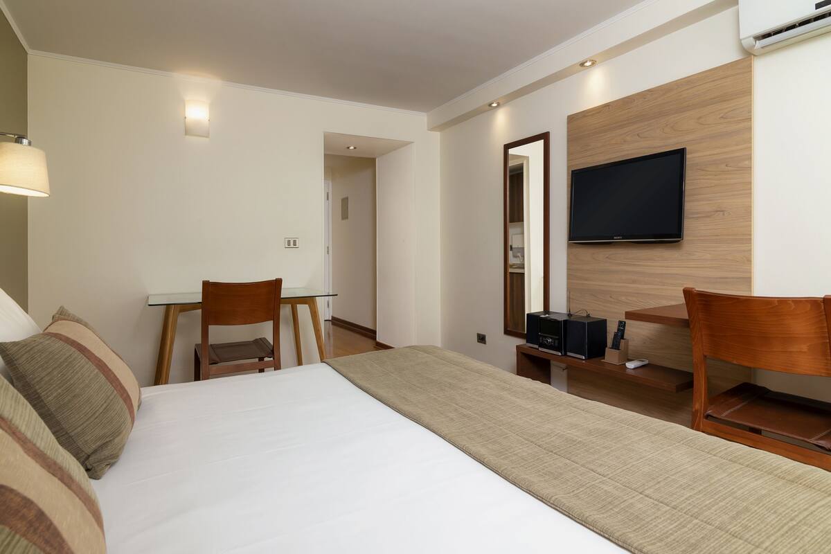 Habitación de hotel con cama, dos sillas de madera, un escritorio, un televisor de pantalla plana montado en una pared de madera y un equipo de aire acondicionado.
