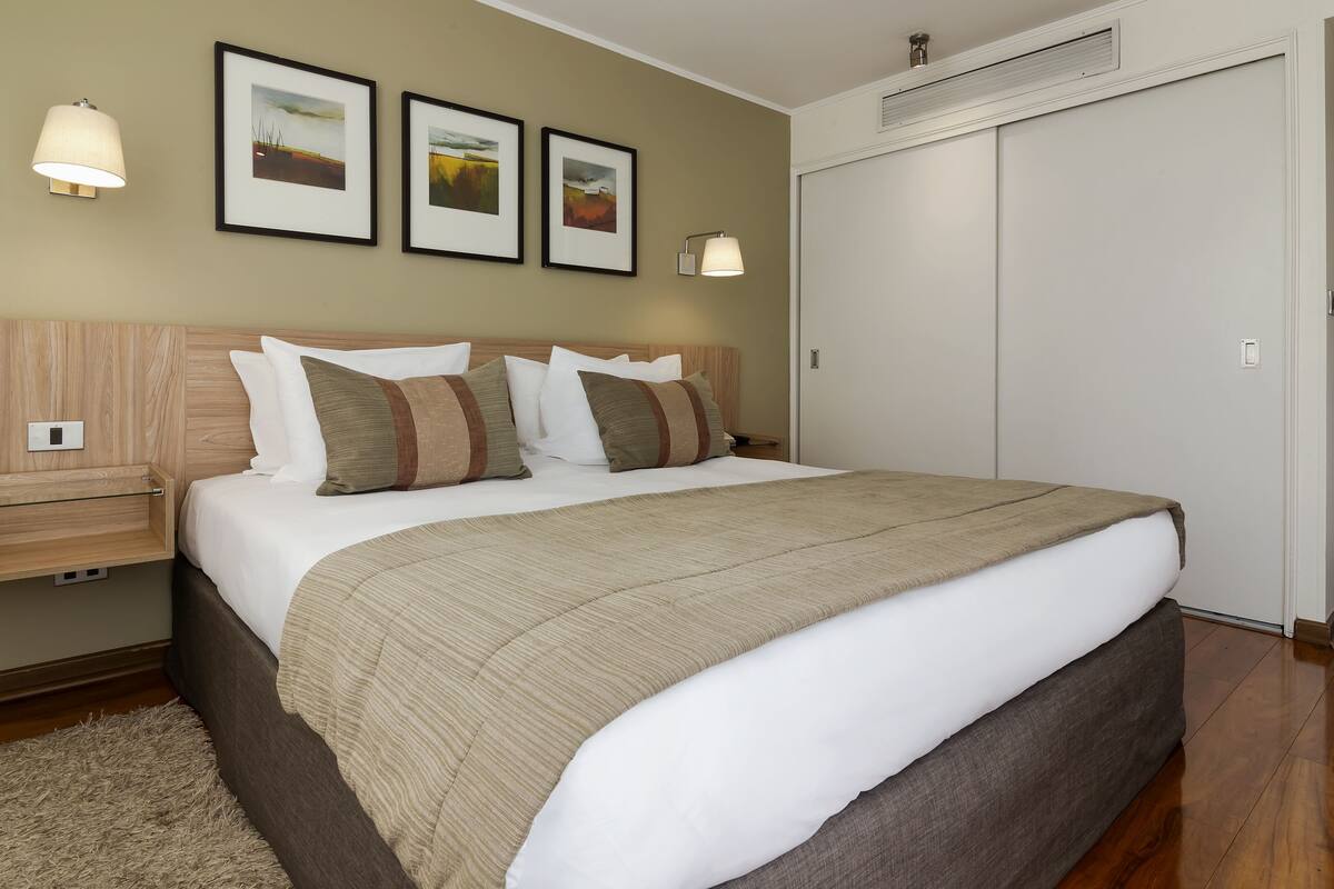 Una cama bien hecha con una colcha color beige, un cabecero de madera y dos lámparas fijadas a la pared.