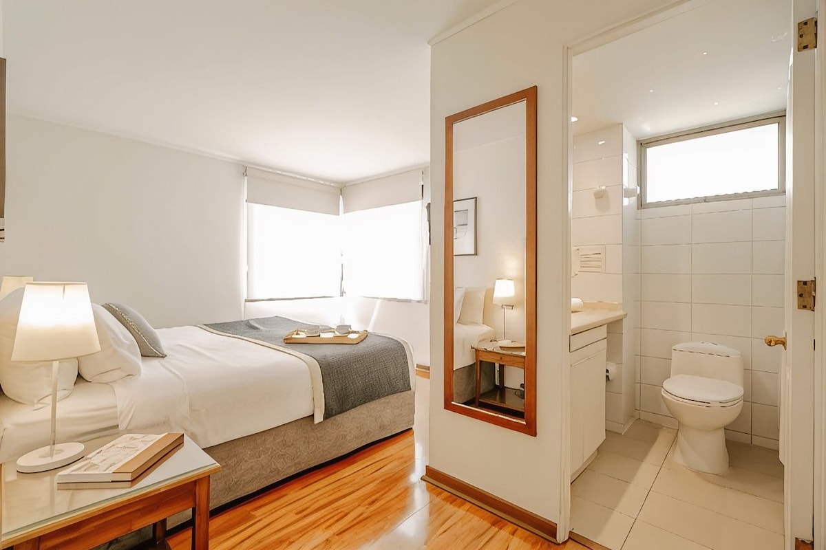 Quarto de hotel com cama, mesa de cabeceira, abajur e espelho.