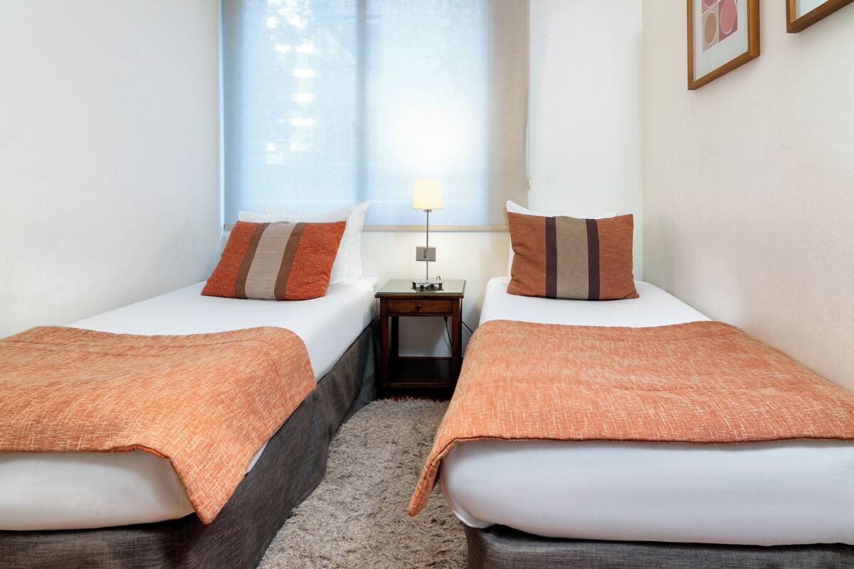 Habitación de hotel con dos camas individuales, cada una con una manta naranja, una mesita de noche con lámpara y una ventana con persianas.