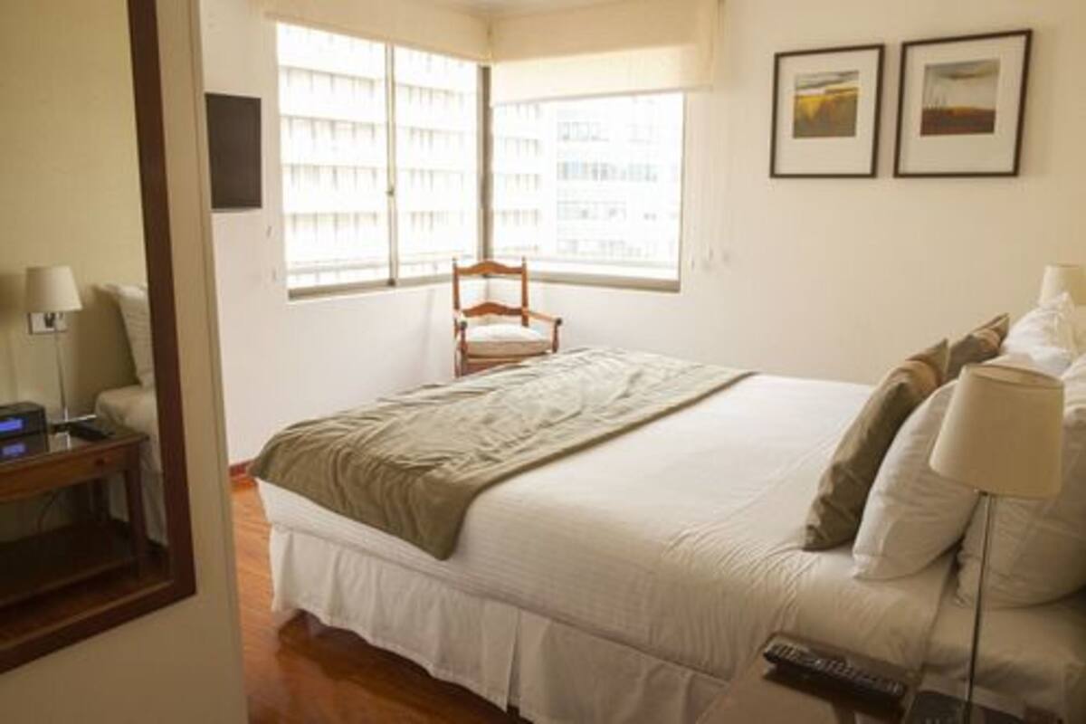 Un dormitorio con una cama, una silla, un escritorio con una lámpara y dos cuadros en la pared.