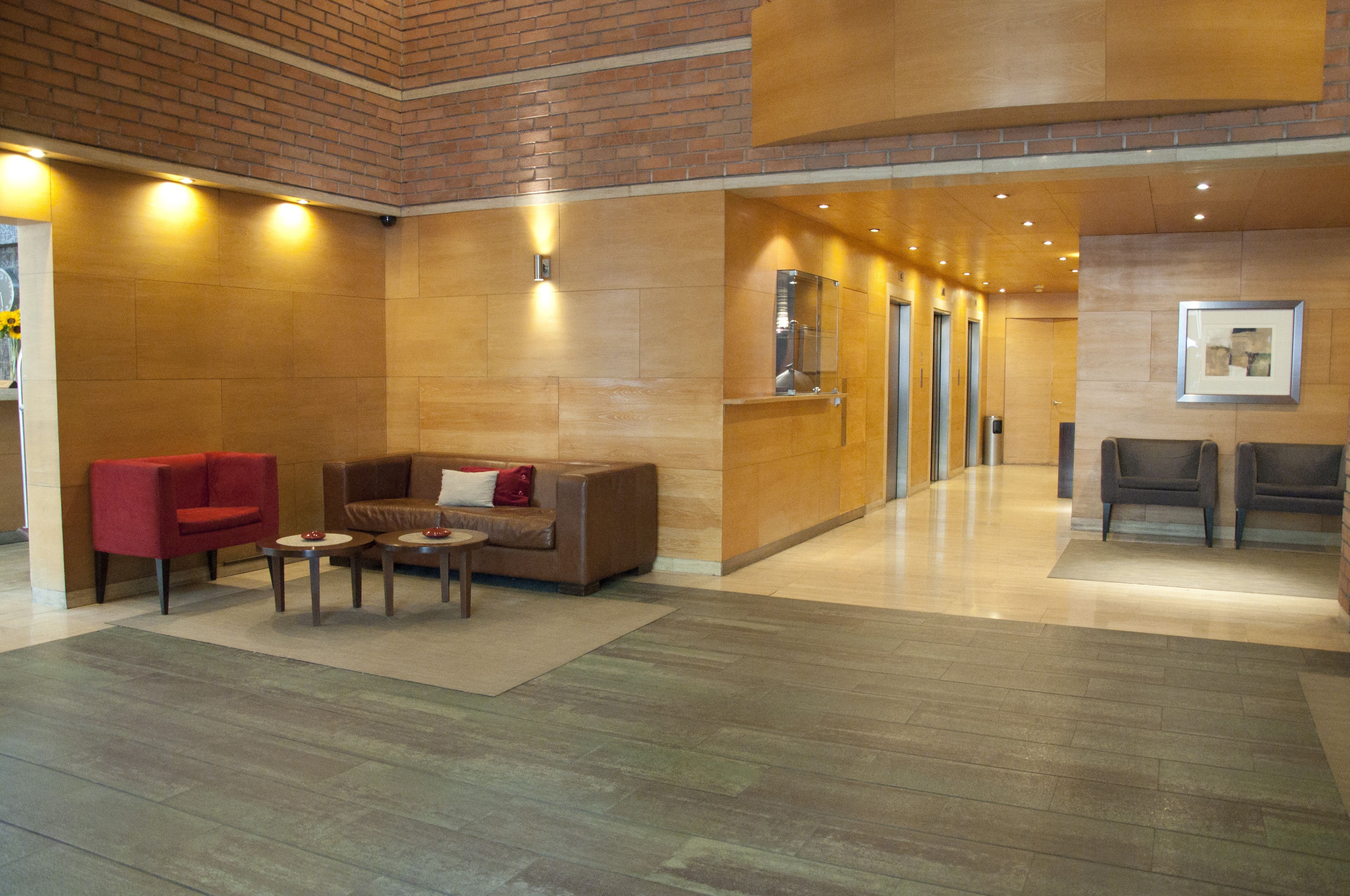 A lobby with a red armchair, a brown sofa, and two armchairs. There are two small round tables with red and white objects on them. The floor is tiled, and the walls are made of wood panels.