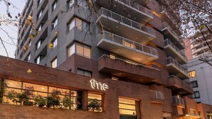 A multi-story hotel with a modern design, featuring balconies on each floor, a ground-level entrance with a sign, and a visible parking area. - Time Suite - Comfort Apartments (Santiago)