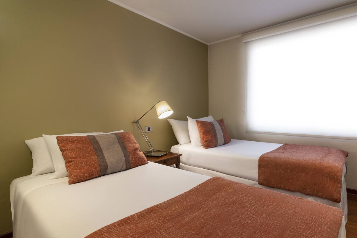 Habitación de hotel con dos camas, una mesita de noche con lámpara y un ventanal que deja entrar luz natural.