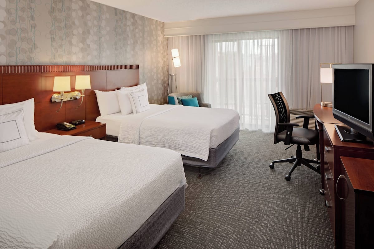 A hotel room with two beds, a desk with a TV, a chair, and a lamp.