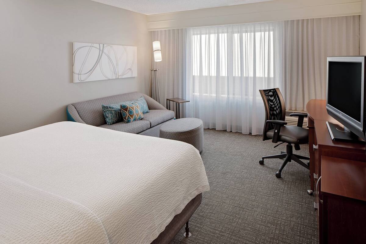 A hotel room with a large bed, a desk with a TV, a chair, a small table, and a window with curtains.