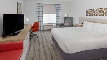 A hotel room with a large bed, a desk with a flat-screen TV, a red chair, a grey armchair, and a window with patterned curtains.