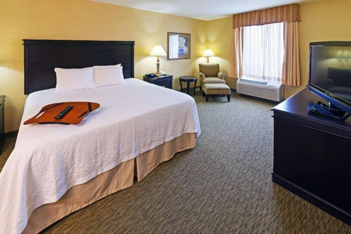 A hotel room with a large bed, a desk with a television, a chair, an air conditioning unit, and a lamp.