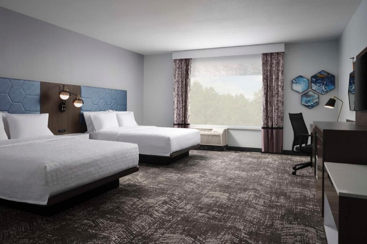 Une chambre d’hôtel avec deux lits, un bureau, une chaise, une fenêtre avec des rideaux et des œuvres d’art accrochées au mur.