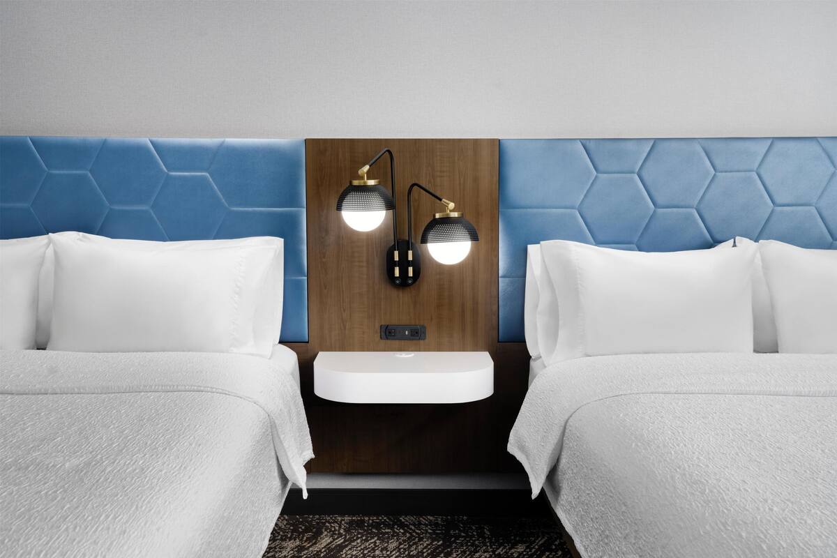 A hotel room with two beds, a nightstand, a wall-mounted lamp, and a small sink.