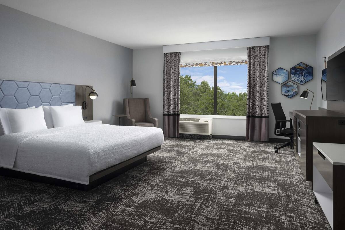 A hotel room with a large bed, a desk, a chair, a lamp, and a view of trees through the window.